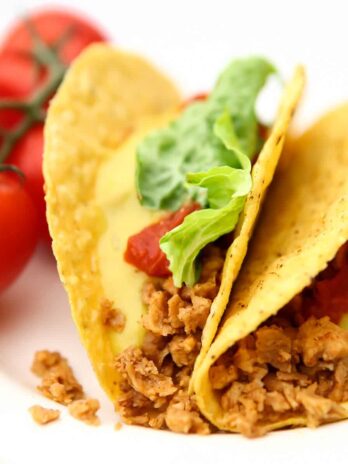 Two vegan tacos filled with tvp taco meat and topped with lettuce, tomatoes, and vegan nacho cheese sauce.