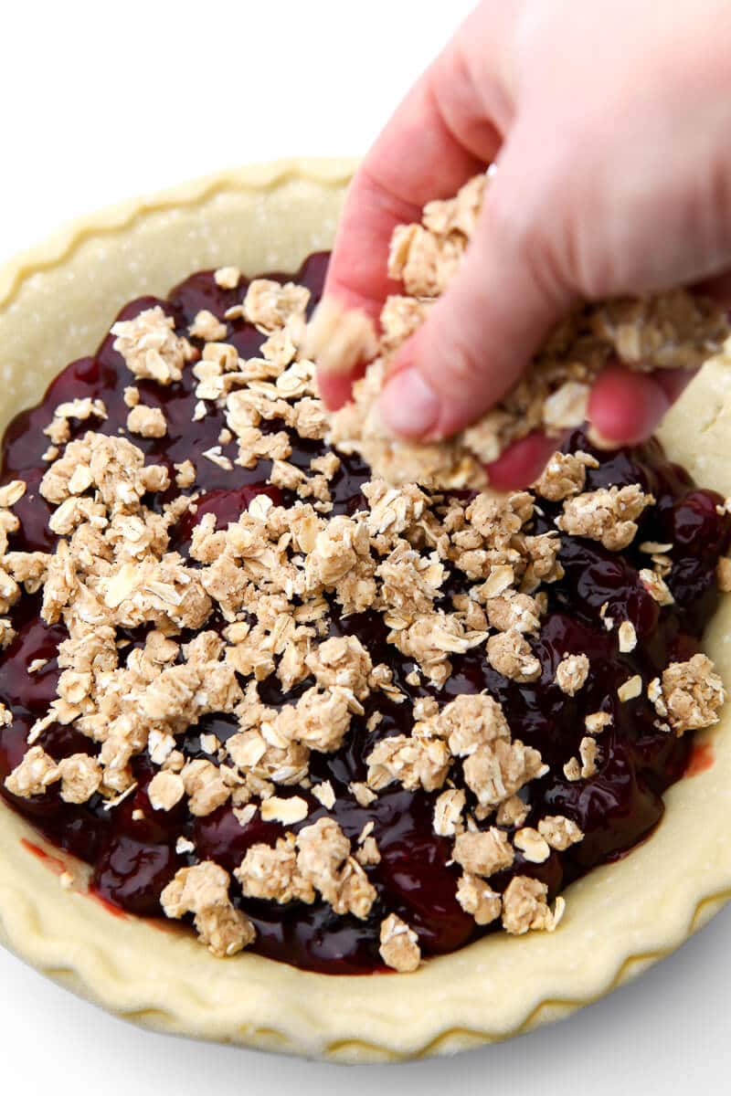 A hand sprinkling crumble topping on a cherry pie before baking.