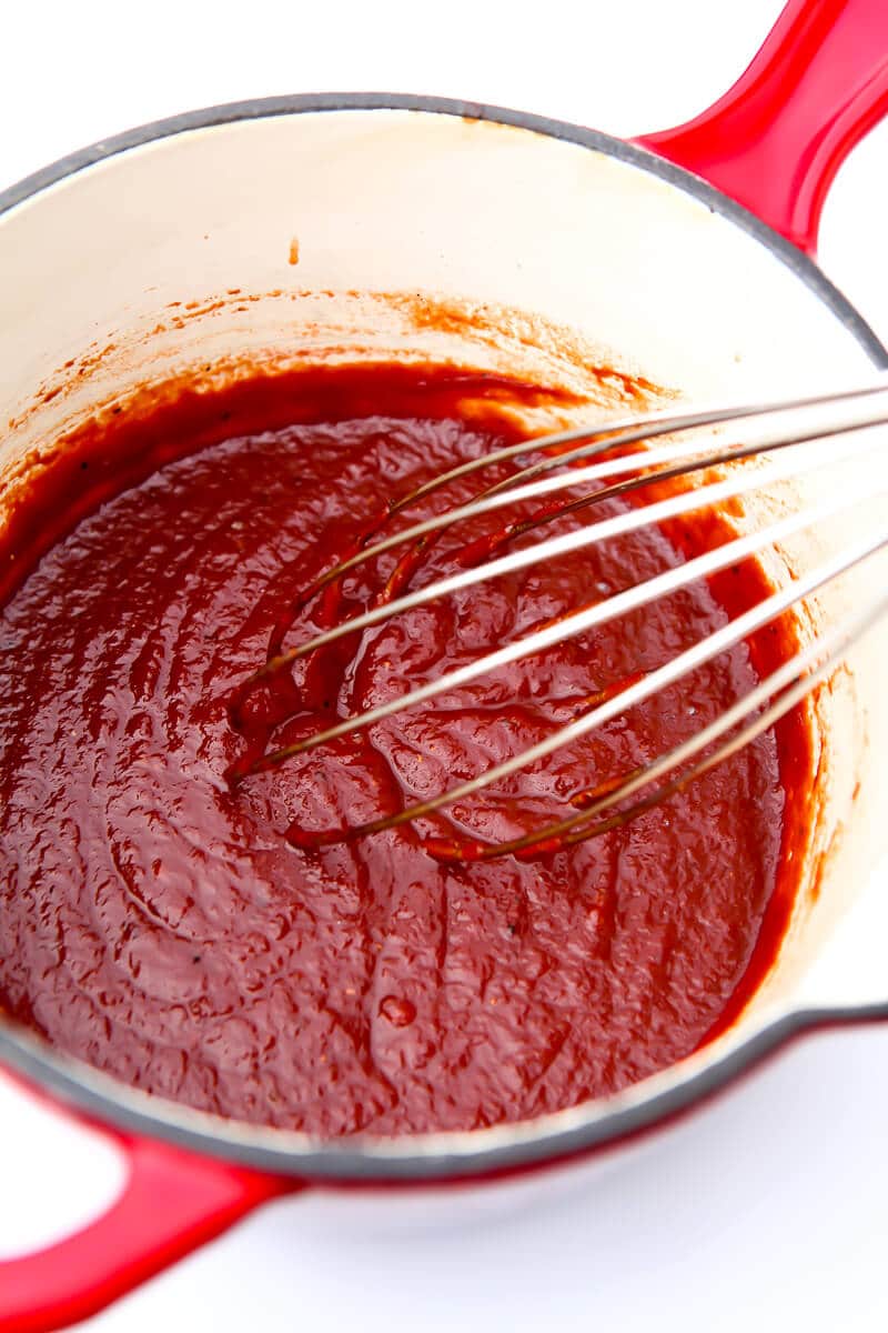 Homemade refined sugar free BBQ sauce in a red saucepan being stirred with a whisk.