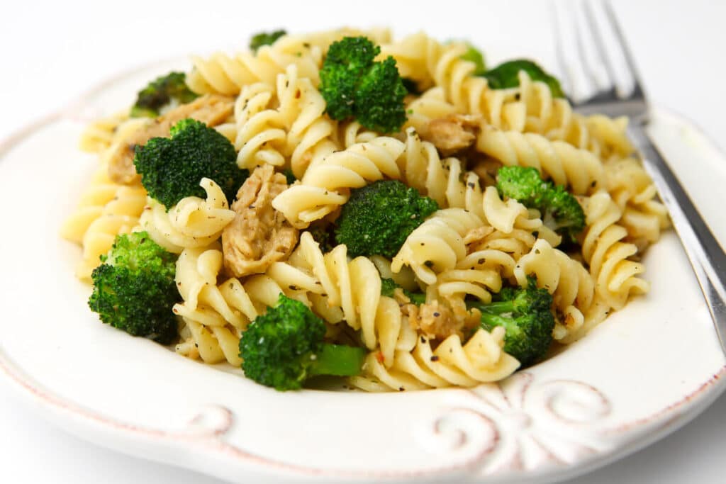 A white plate filled with broccoli pasta with vegan chicken with a fork on the side.