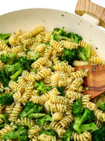 A large white wok filled with broccoli tossed with pasta with a wooden spoon stirring it.
