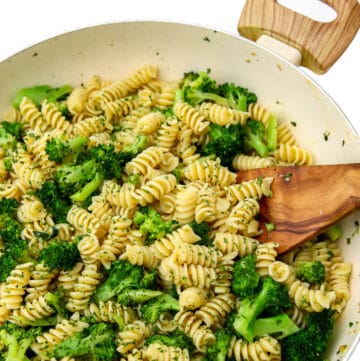 A large white wok filled with broccoli tossed with pasta with a wooden spoon stirring it.