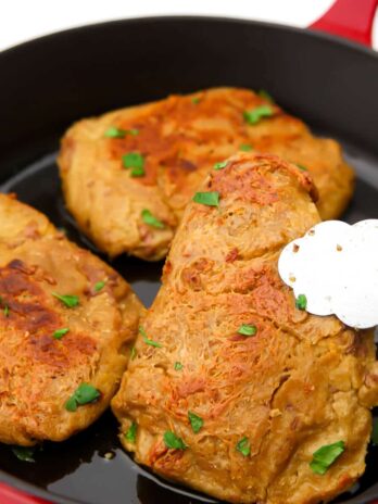Three vegan chicken breast being sauted in a red cast iron frying pan and a piece held up with tongs.