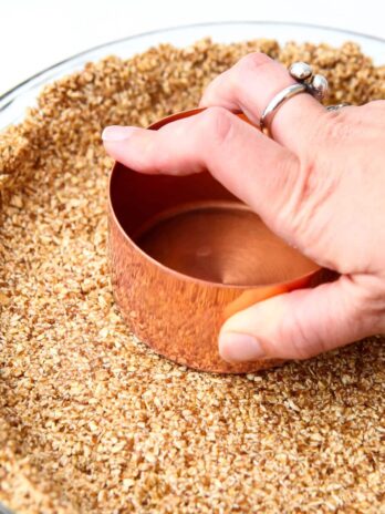 A gluten-free vegan graham cracker crust being packed down with the bottom of a copper measuring cup.