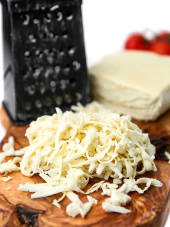 A block of vegan mozzarella cheese on a cutting board with a cheese grater and some of it shredded.