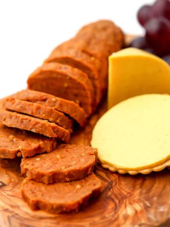 Homemade vegan pepperoni on a wooden cutting board next to sliced cheese and purple grapes.
