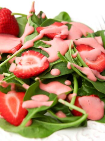 A white plate full of baby spinach and sliced strawberries with pink strawberry dressing drizzled over it.