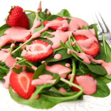 A white plate full of baby spinach and sliced strawberries with pink strawberry dressing drizzled over it.