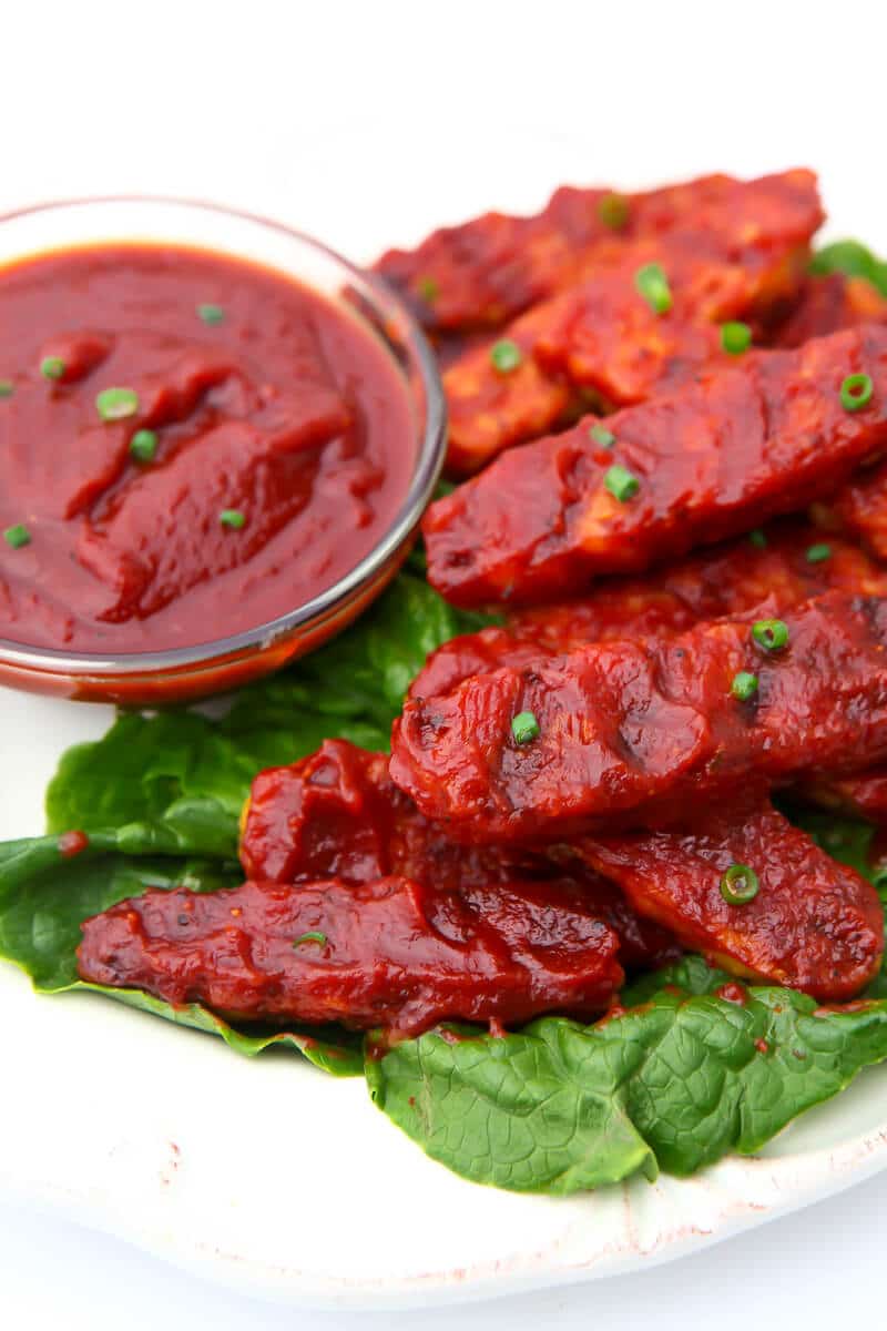 A plate of bbq tempeh on a bed of lettuce with barbeque sauce on the side.