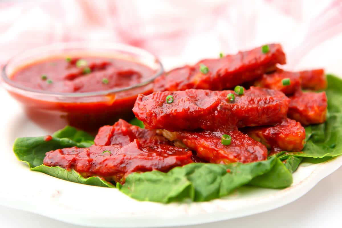 A plate of barbeque tempeh on a bed of lettuce with a bowl of BBQ sauce on the side.