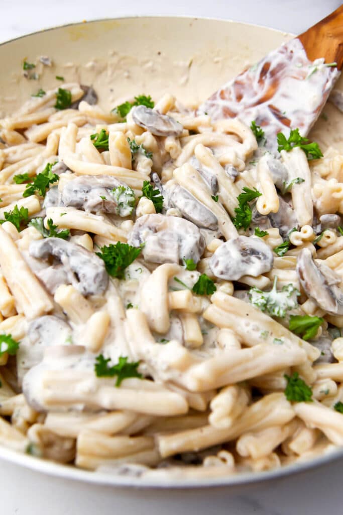 A creamy mushroom pasta in a large wok being stirred with a wooden spoon.