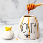 A white honey pot filled with vegan honey with some dripping down the sides.