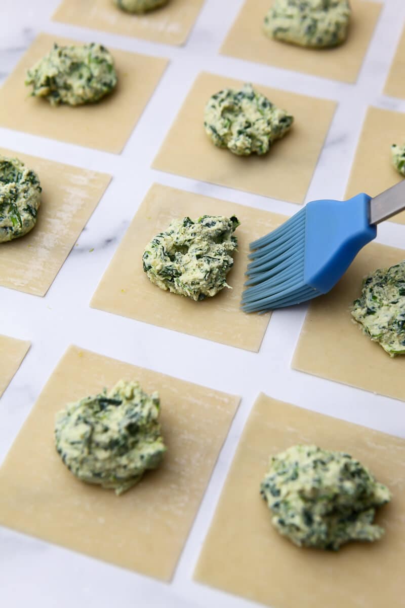 A blue pastry brush wetting the sides of the ravioli to seal them up.