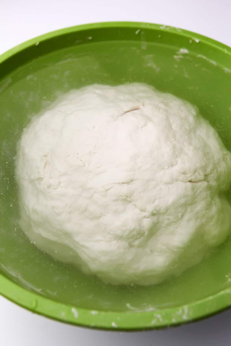 A ball of flour and water dough in a large green bowl covered with water.