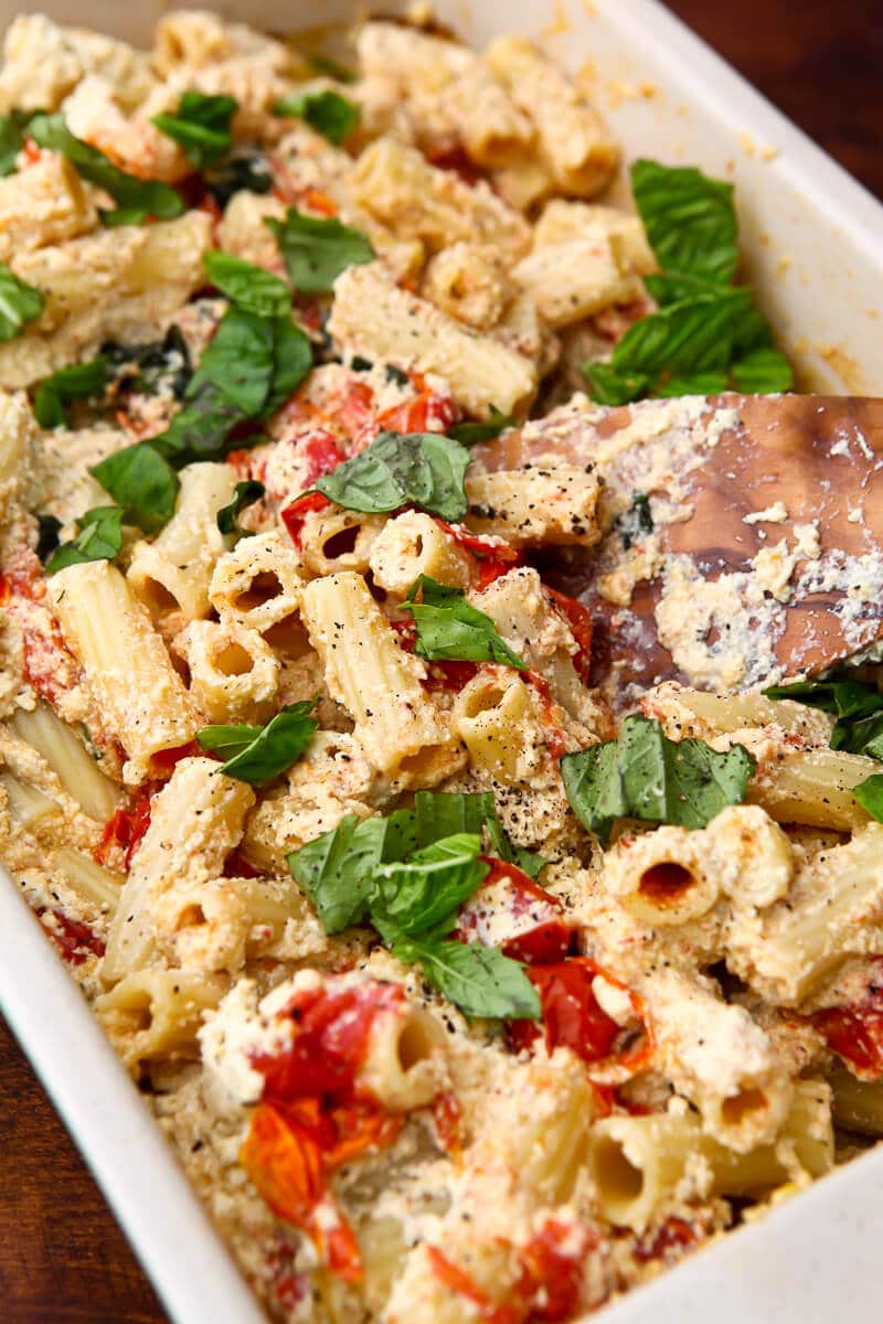 A casserole dish filled with baked vegan feta, cherry tomatoes, and pasta mixed together with fresh basil on top.