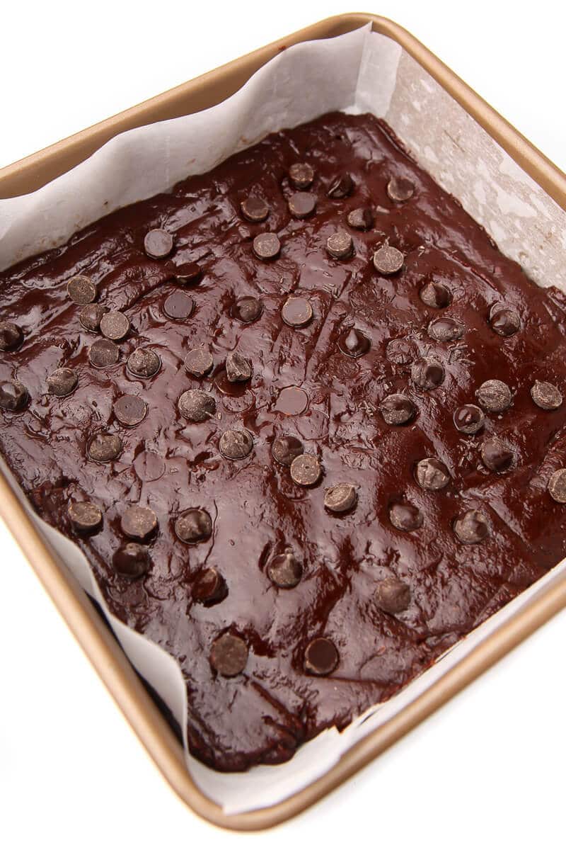 Vegan brownie batter pressing in a pan that has been lined with parchment paper and topped with chocolate chips.