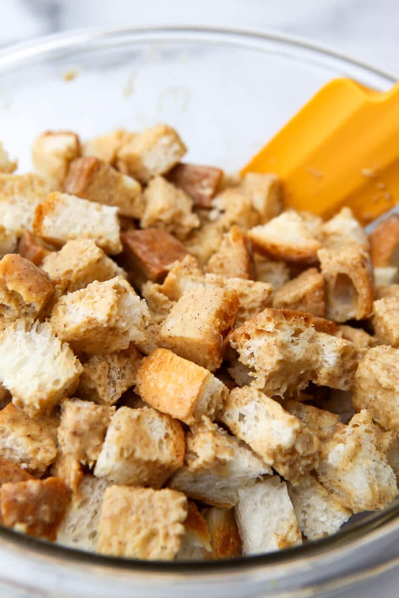 A bowl of bread cubes with vegan French toast custard being stirred with  a rubber spatula.