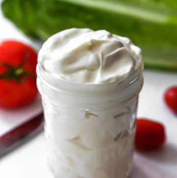 A mason jar filled with vegan mayo made with soy milk and oil with some lettuce and tomatoes around it.