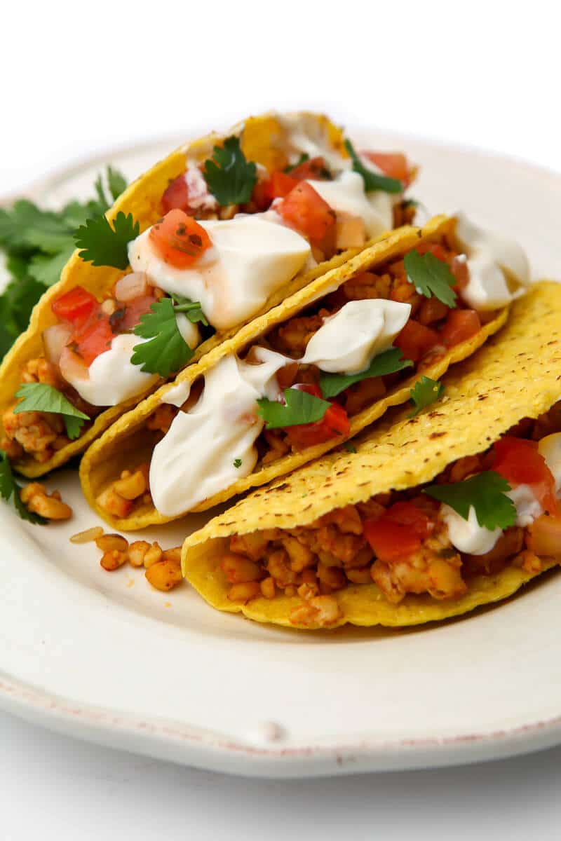 Three tacos on a white plate filled with  tempeh taco meat, salsa, and vegan sour cream.