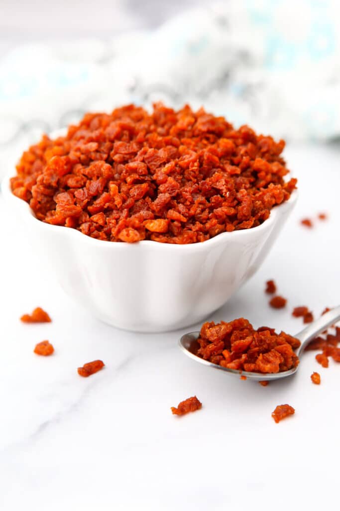 A white bowl filled with bacon bits made from TVP and spices with a spoon on the side.