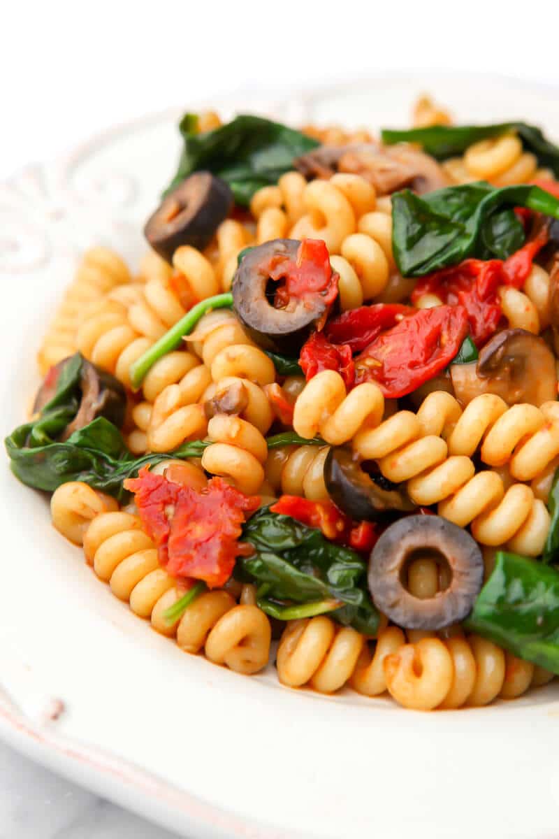 Pasta salad with tomatoes, mushrooms, black olives, spinach, and corkscrew pasta on a white plate.