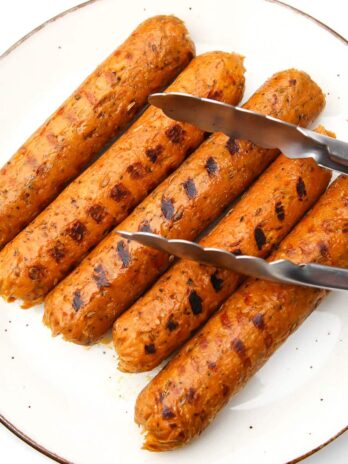 Five vegan Italian sausages on a plate after being grilled with tongs on them.
