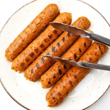 Five vegan Italian sausages on a plate after being grilled with tongs on them.