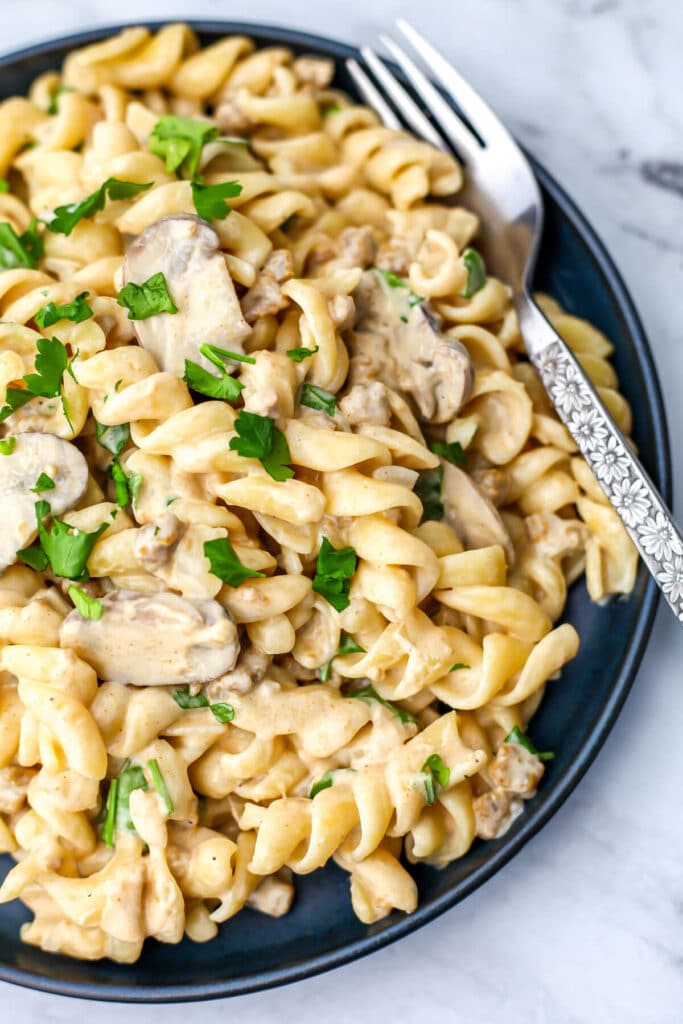 A top view of a creamy vegan stroganoff made with vegan sour cream, mushrooms, and vegan beef crumbles served over thick spiral nood.es with a fork on the side.
