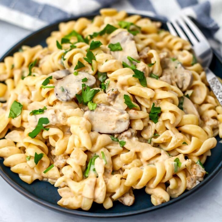Vegan beef stroganoff over spiral noodles on a blue plate with a fork on the side.
