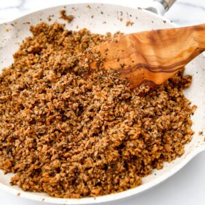 A white skillet filled with homemade vegan ground beef being stirred with a wooden spoon.