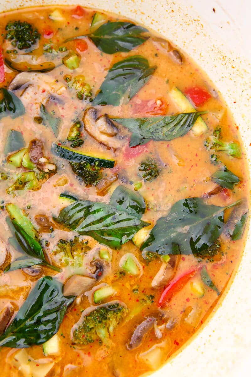 A pot full of vegan red curry sauce with red peppers, broccoli, mushrooms, pineapple and basil.