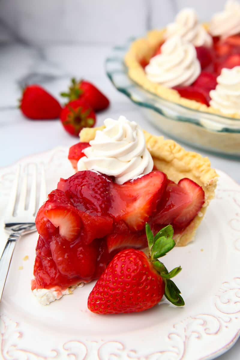 A slice of strawberry pie served on a white plate with a full pie behind it.