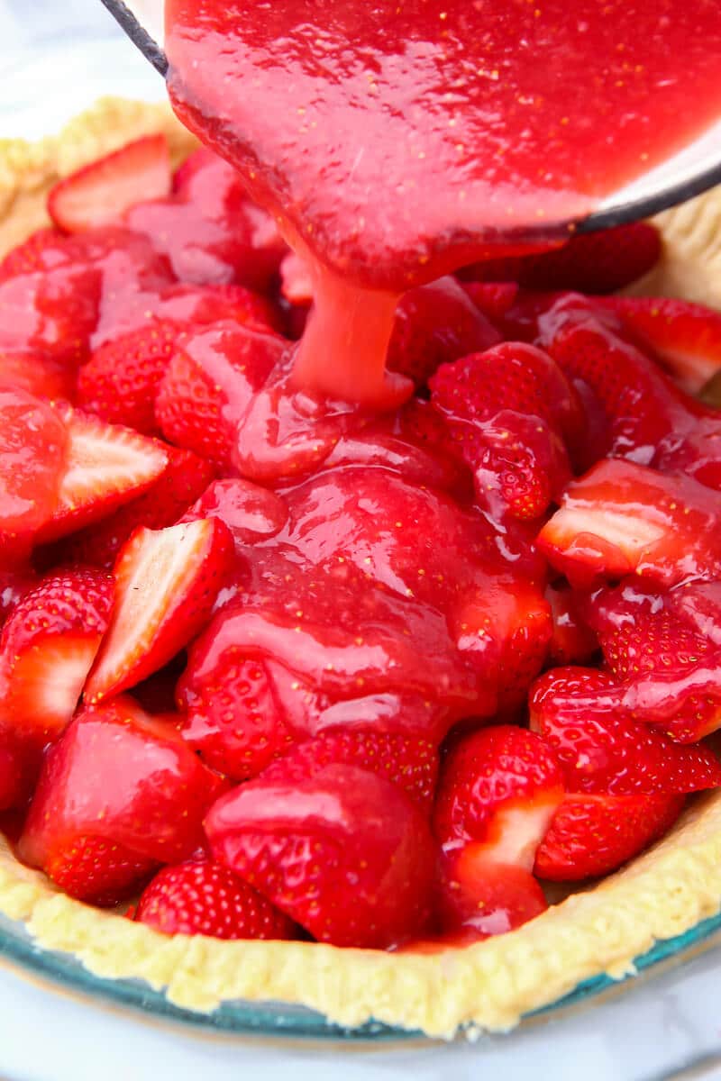 Vegan strawberry sauce being poured over strawberries in a pre-baked pie shell.
