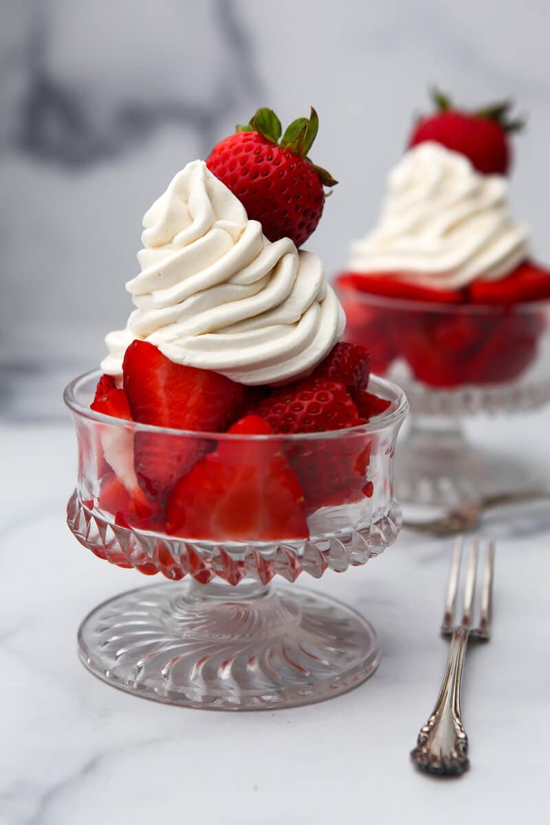 Two glass dishes filled with strawberries topped with dairy free whipped cream and another strawberry on top of that.