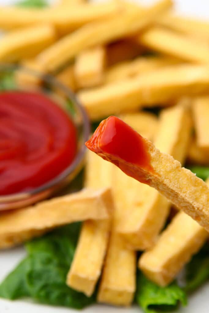 A close up of a French fry made of tofu dipped in ketchup.