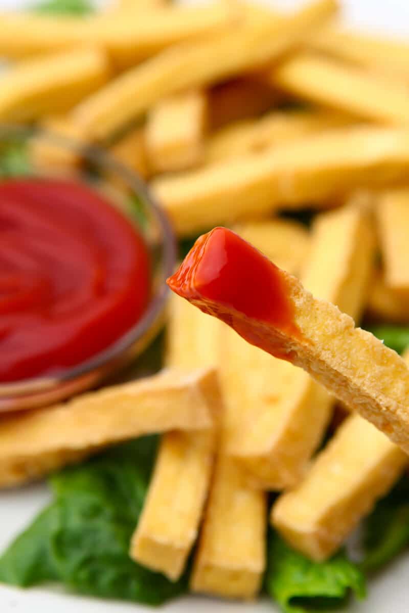 A close up of a French fry made of tofu dipped in ketchup.
