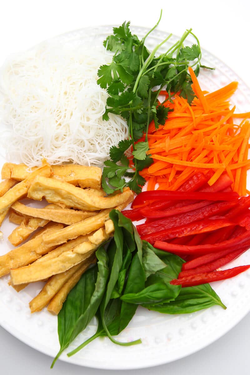 The ingredients to make tofu spring rolls on a white plate including basil, carrots, red pepper, cilantro, and rice noodles.