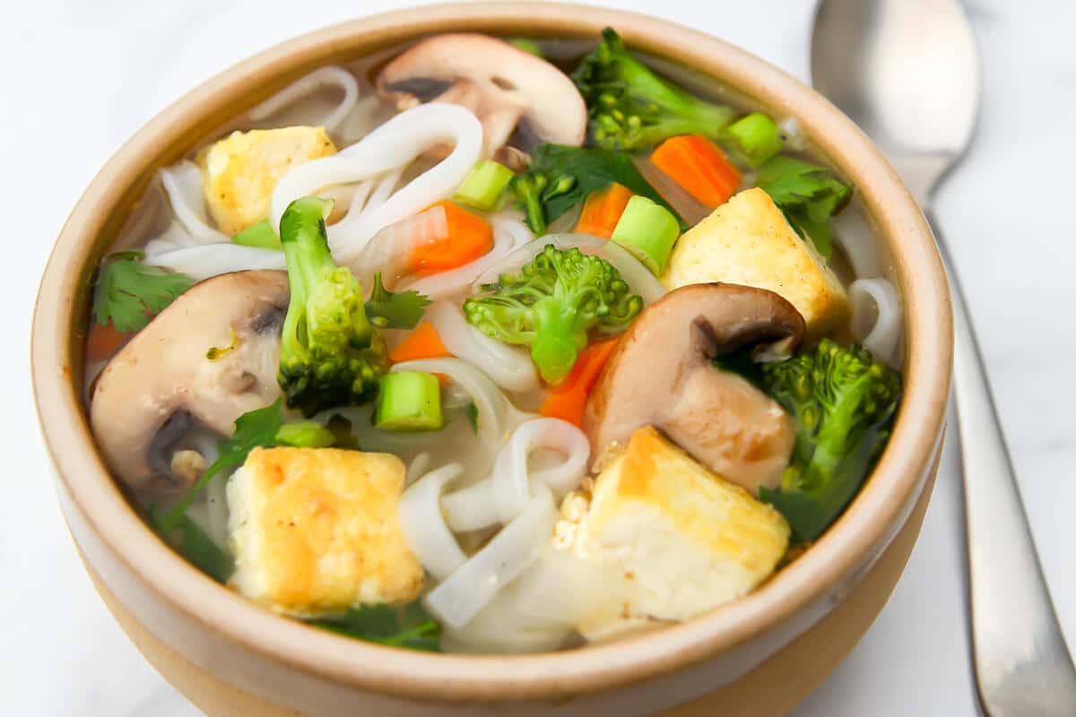 A bowl of vegan pho soup with veggies, tofu, and rice noodles with a spoon on the side.