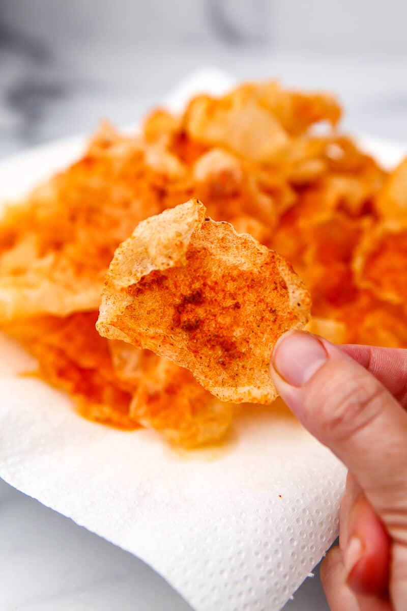 A hand holding a piece of fried rice paper seasoned to taste like pork rinds with a plate full behind it.
