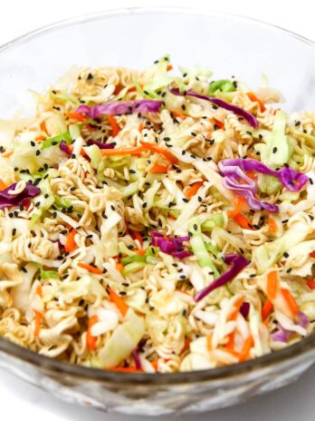 A glass bowl filled with vegan ramen noodle salad with cabbage and carrots and sprinkled with black sesame seeds.