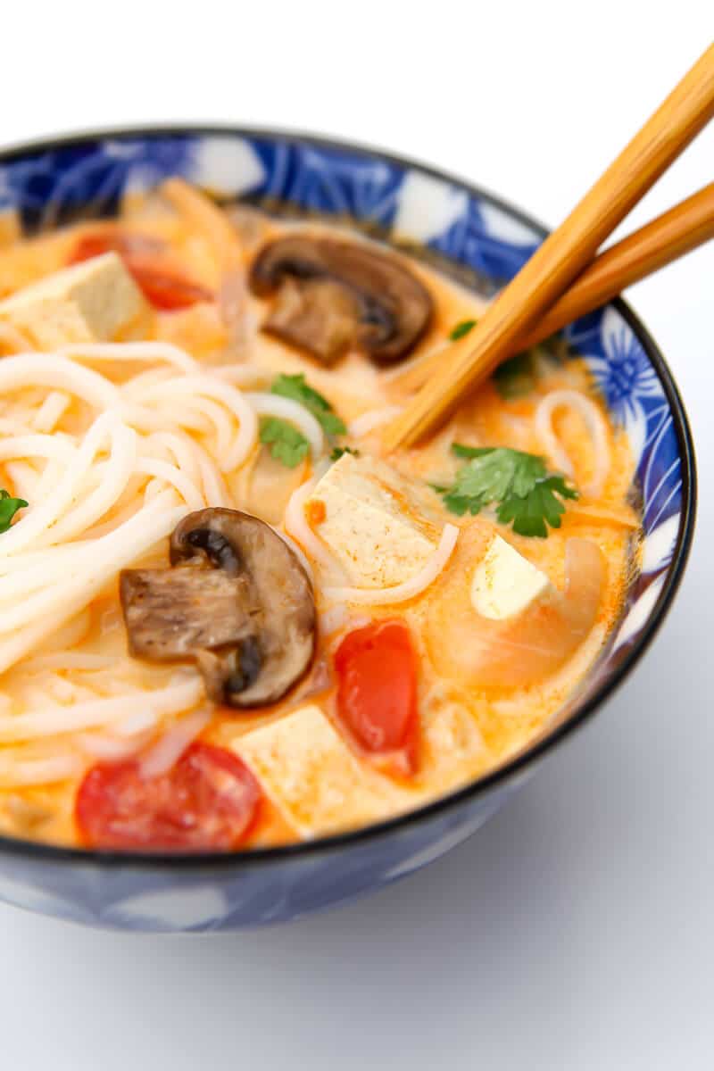 A bowl of tom kha soup with tofu, mushrooms, tomatoes, and rice noodles with chop sticks in it.