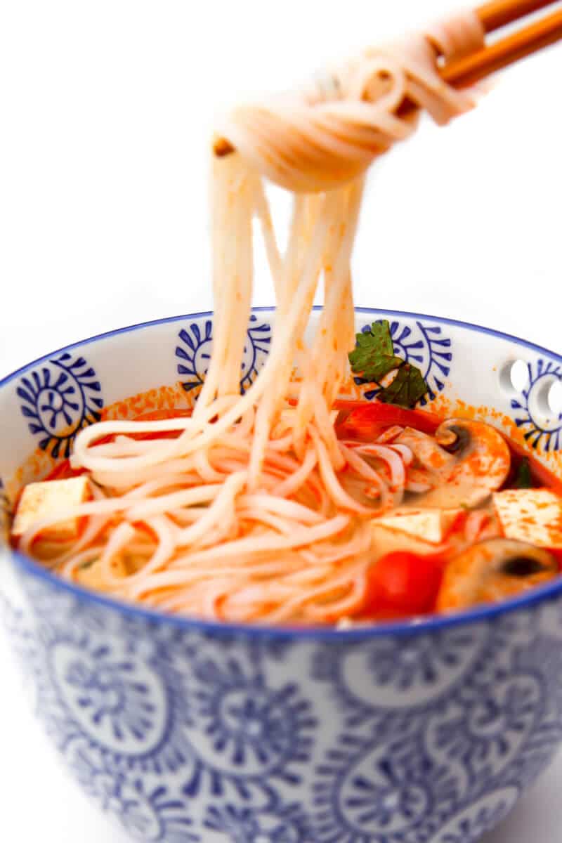 A blue bowl of vegan tom yum soup with chopsticks pulling up some rice noodles. 