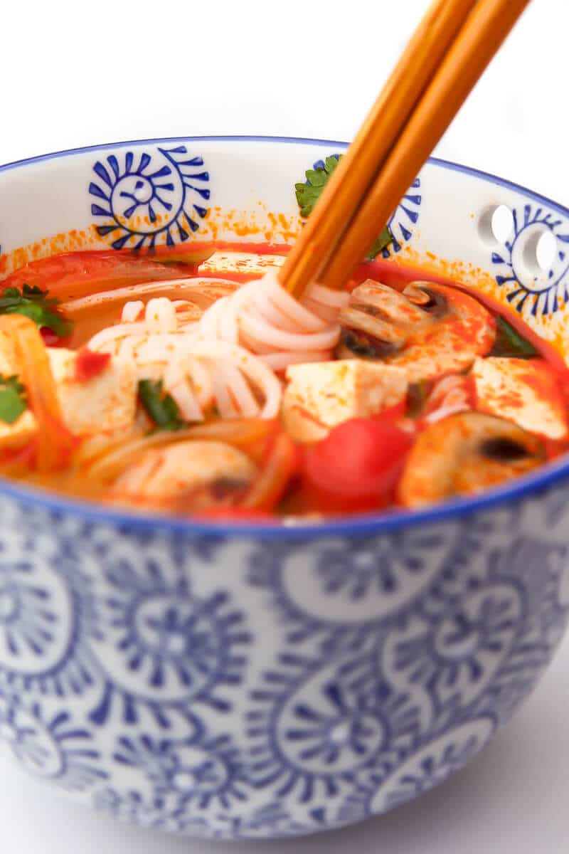 A blue bowl filled with tom yum soup and rice noodles with chop sticks in it.