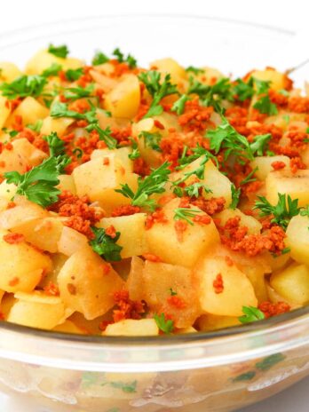 A large glass bowl filled with vegan German potato salad with bacon bits and parsley on top.