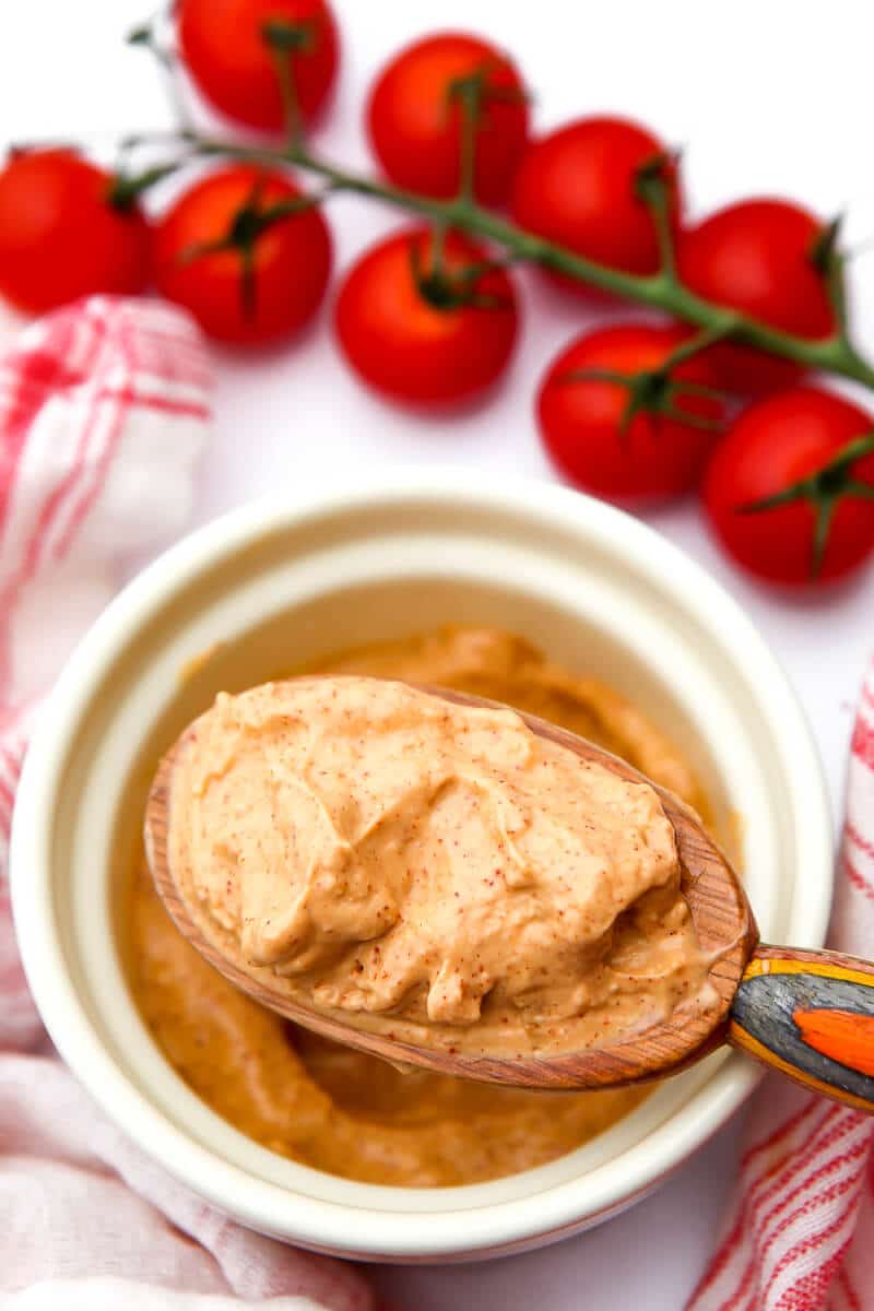 A spoon full of thick homemade vegan chipotle mayo with cherry tomatoes next to a bowl.