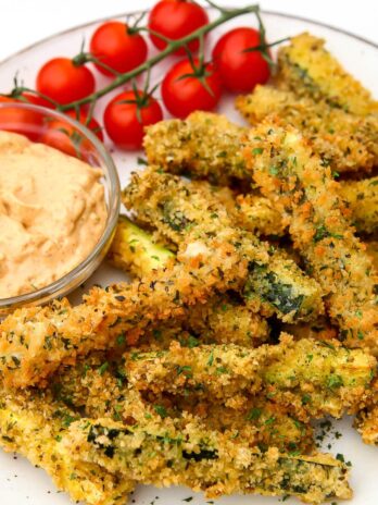 A plate full of deep fried breaded zucchini fries with tomatoes and dipping sauce on the side.
