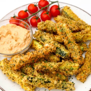A plate full of deep fried breaded zucchini fries with tomatoes and dipping sauce on the side.