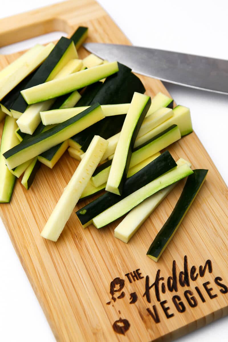 A zucchini cut into French fry shaped pieces on a cutting board with a knife on the side.