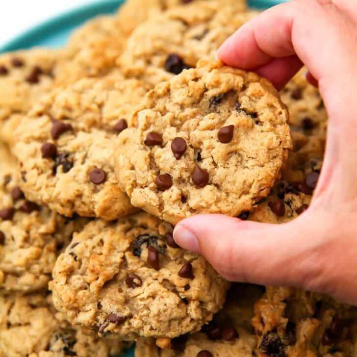 A pile of vegan oatmeal cookies on a blue plate with a hand taking one.