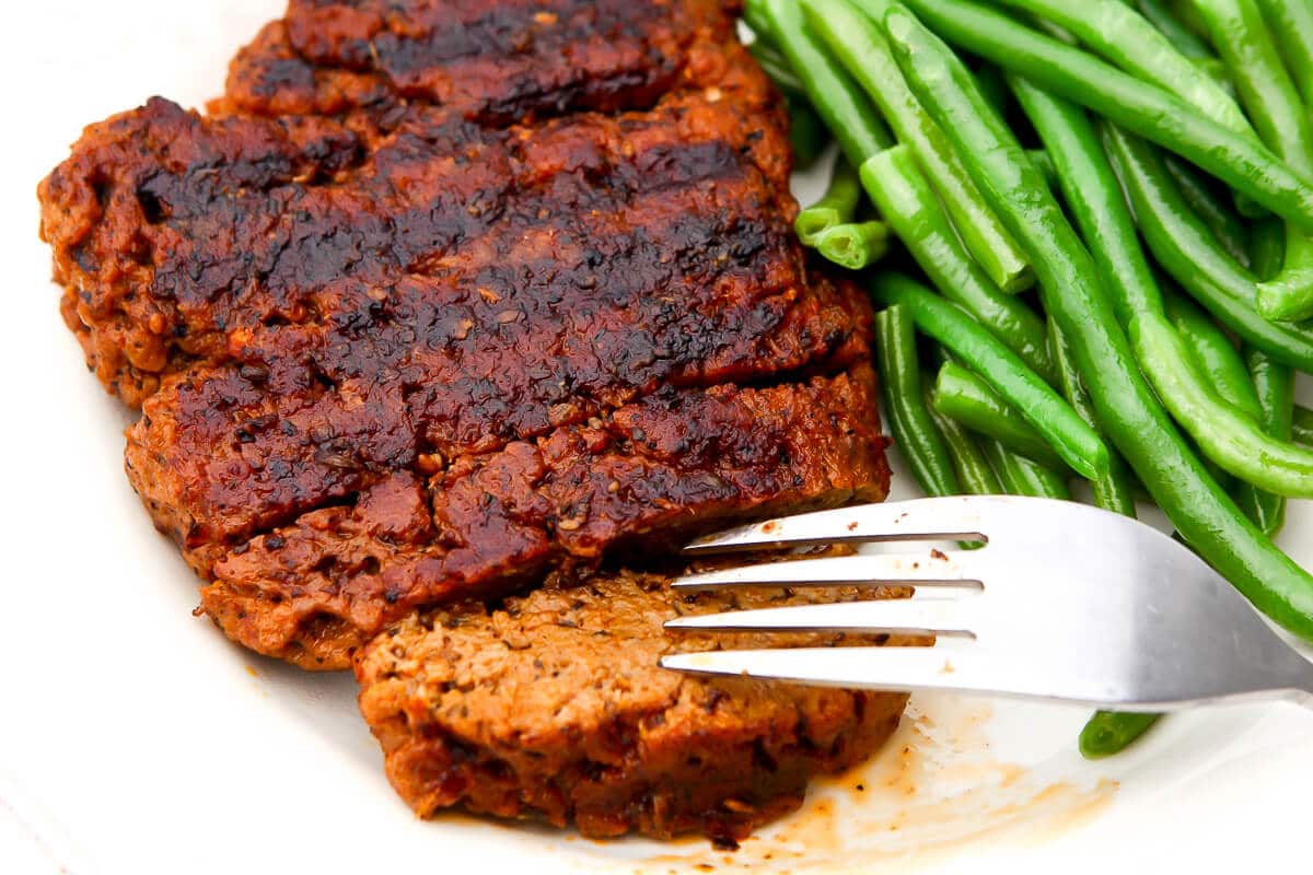 A vegan steak on a white plate with a slice cut off of it and green beans on the side.